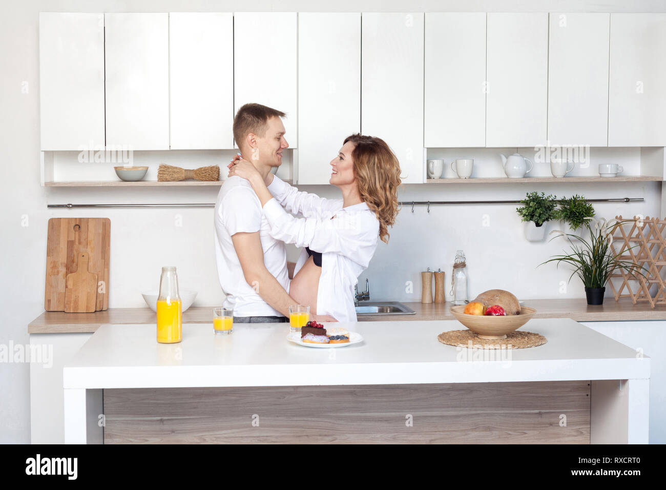 Young married couple embraces standing near table in kitchen. Husband hugs his pregnant wife, putting his hands on her big belly, parents looking to e Stock Photo