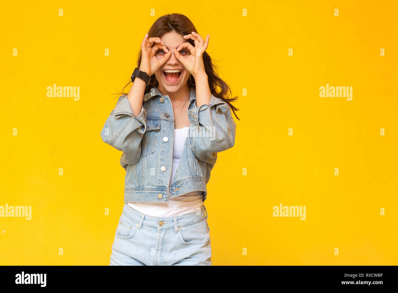 Portrait of surprised beautiful brunette young woman in denim casual style standing and looking at camera with binoculars hands gesture on eyes. indoo Stock Photo