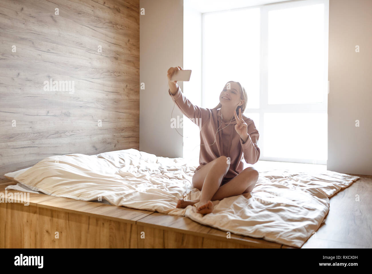 Young beautiful blonde woman sit on bed this morning. She pose on camera and take selfie. Positive model show piece symbol and smile. Earphones in ear Stock Photo