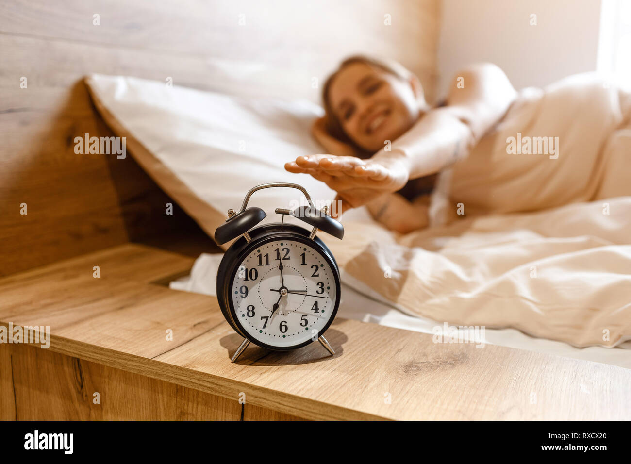 Young beautiful blonde woman lying in bed in morning. Positive happy model reaching alarm clock with hand. . Daylight. Good mood Stock Photo