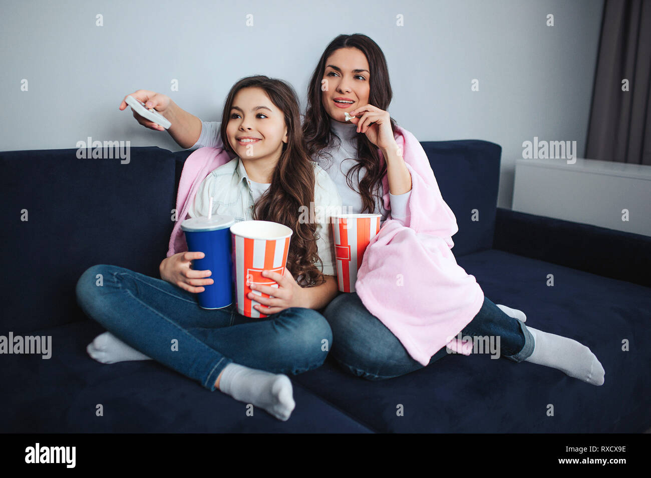 Beautiful brunette caucasian mother and daughter sit together in room. They watch movie. People hold popcorn baskets and cola cup. Adult has remote Stock Photo