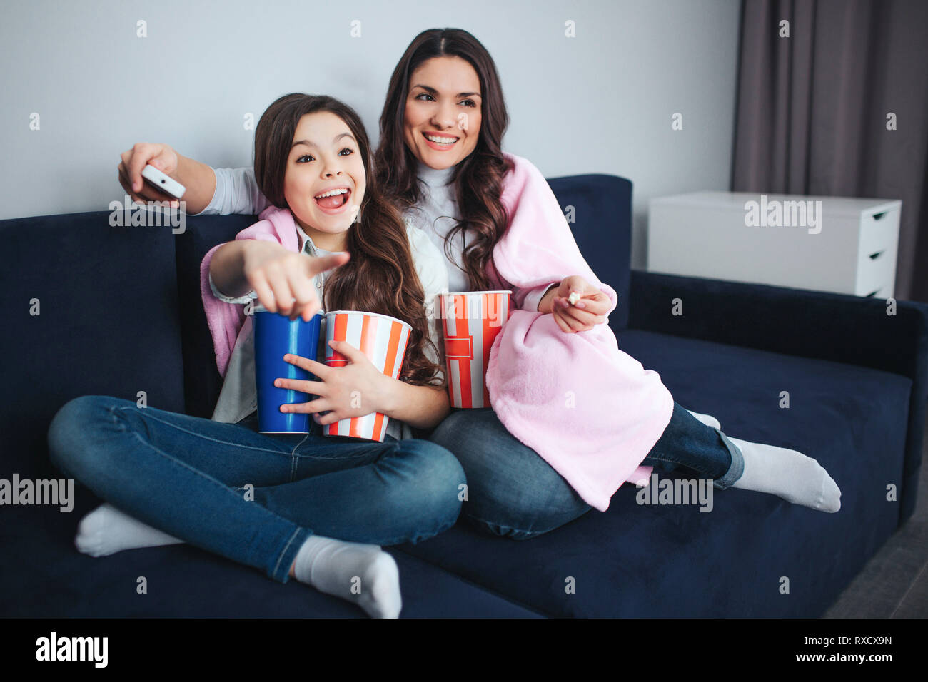 Beautiful brunette caucasian mother and daughter sit together in room. Emotional excited girl point forward. She hold basket popcorn and coke. Mother Stock Photo