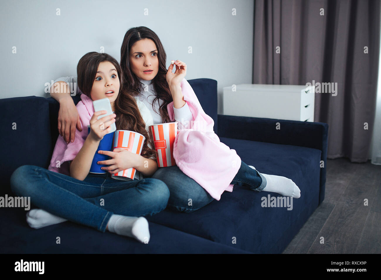 Beautiful brunette caucasian mother and daughter sit together in room. Scared adult and small women watch movie and eat popcorn. Frightened. Stock Photo