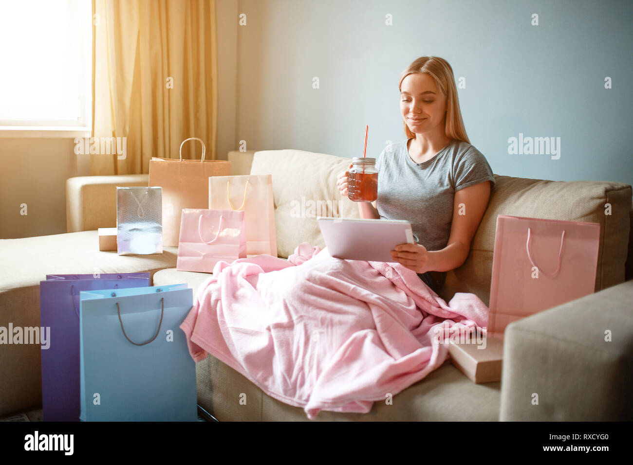 Online shopping at home. Young blonde woman with tea is looking at tablet and choosing goods in online shop while sitting with blanket on a sofa Stock Photo
