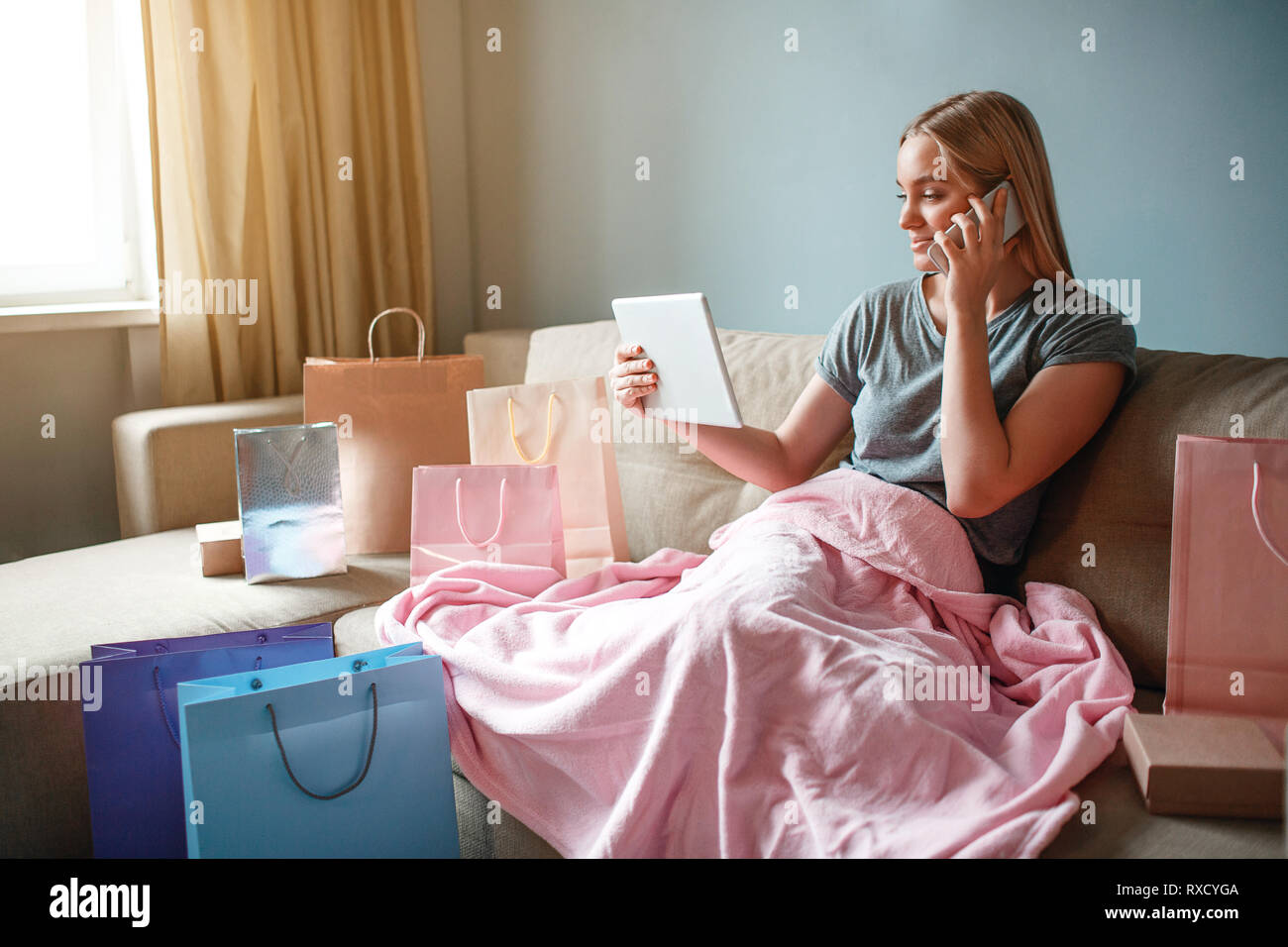 Online shopping at home. Young confident shopper with tablet is ordering by smartphone in online shop while sitting with blanket on a sofa Stock Photo