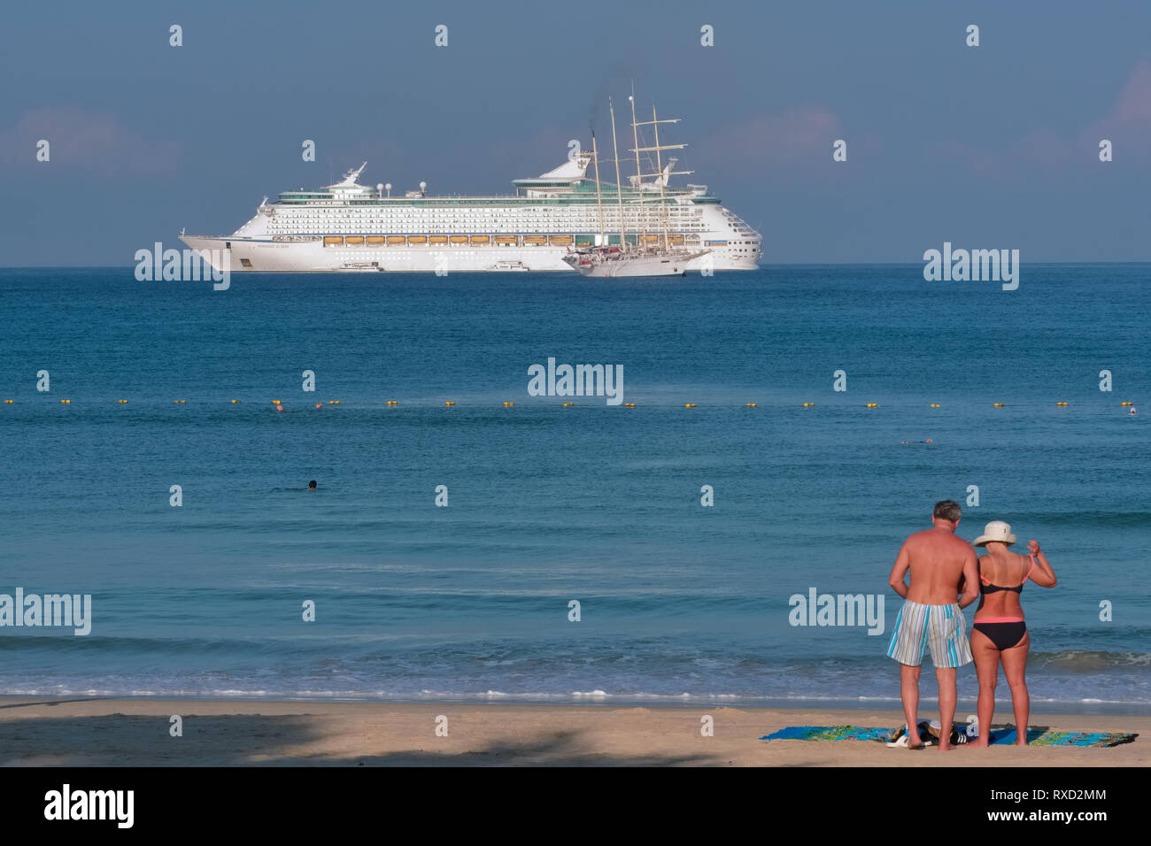 On an early morning, a Western tourist couple stands on Patong Beach, Phuket, Thailand, as a large cruise ship is anchored off the coast Stock Photo