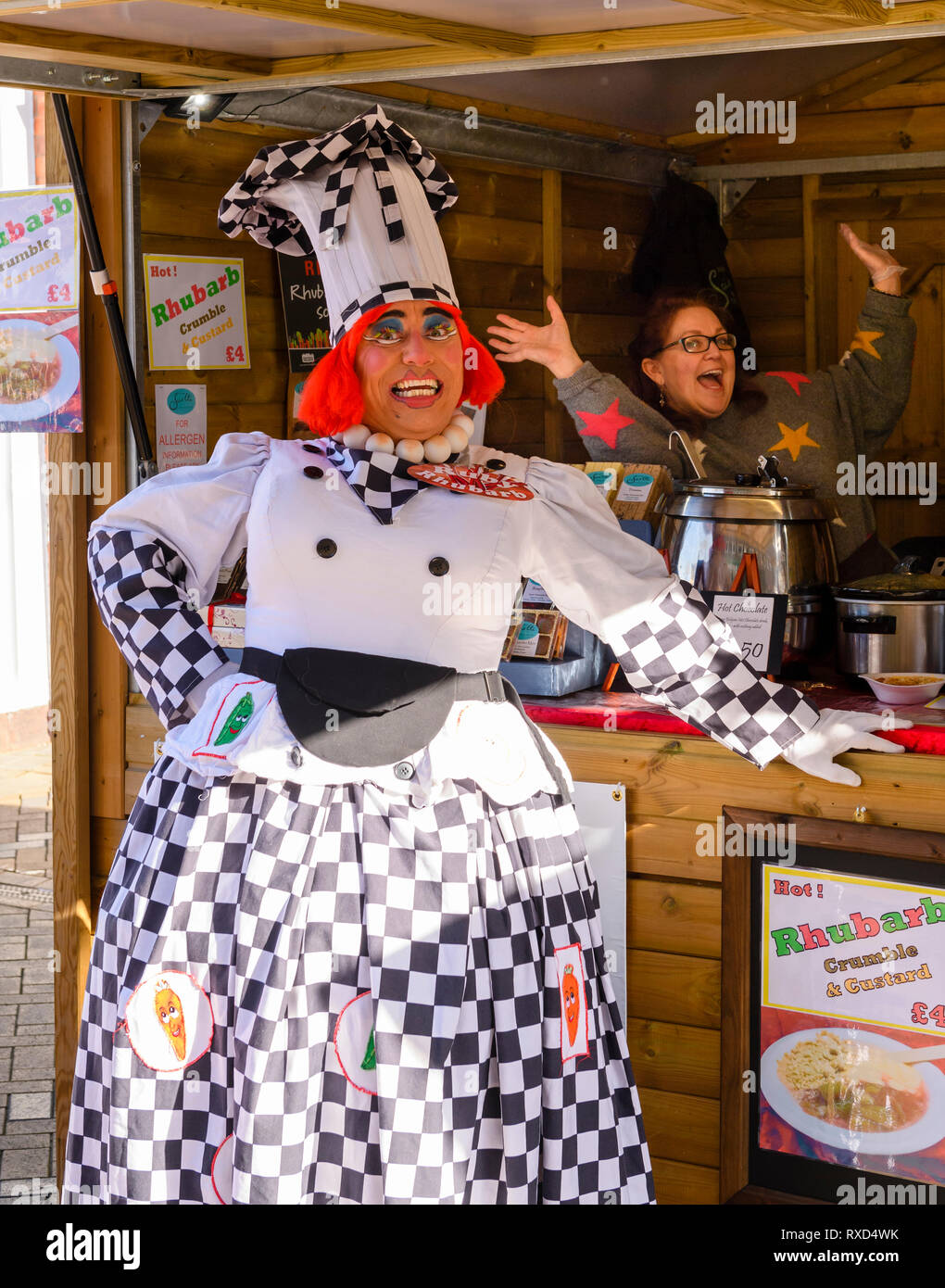 Dame Ruby Rhubarb (comical chef's outfit) & stall trader having fun, smiling & standing by trade stand - Wakefield Food, Drink & Rhubarb Festival 2019 Stock Photo