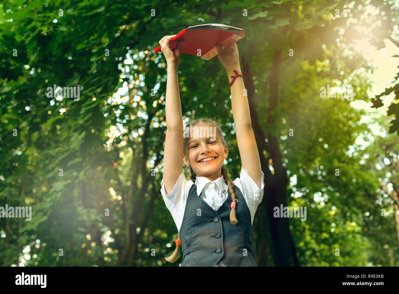 student schoolgirl happy with pigtails in uniform holding books in hands over head in bright sunny day Stock Photo