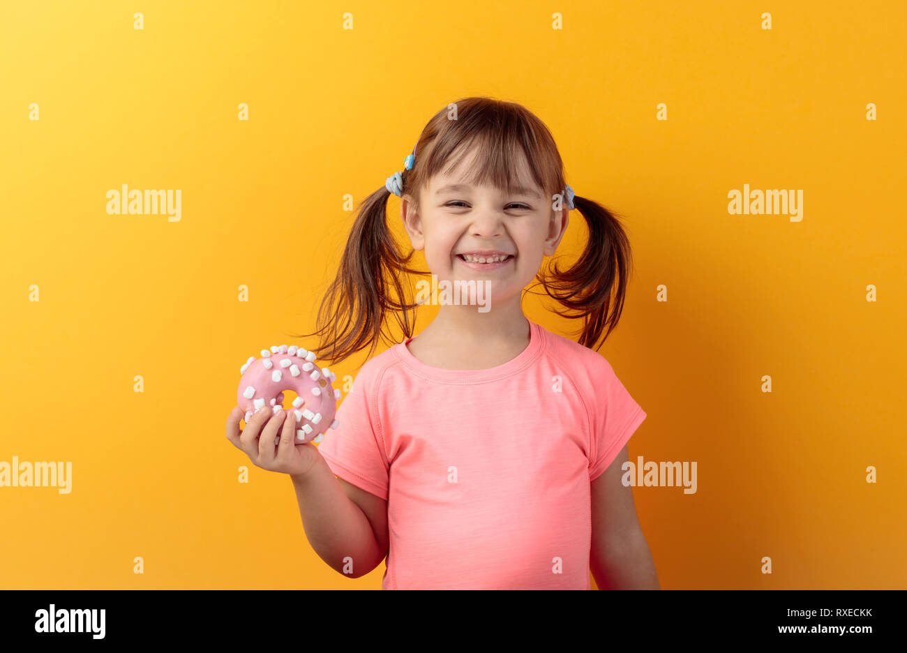 Four-year-old girl in a pink t-shirt eat donut. The girl's hair is tied in tails. Orange background. Stock Photo