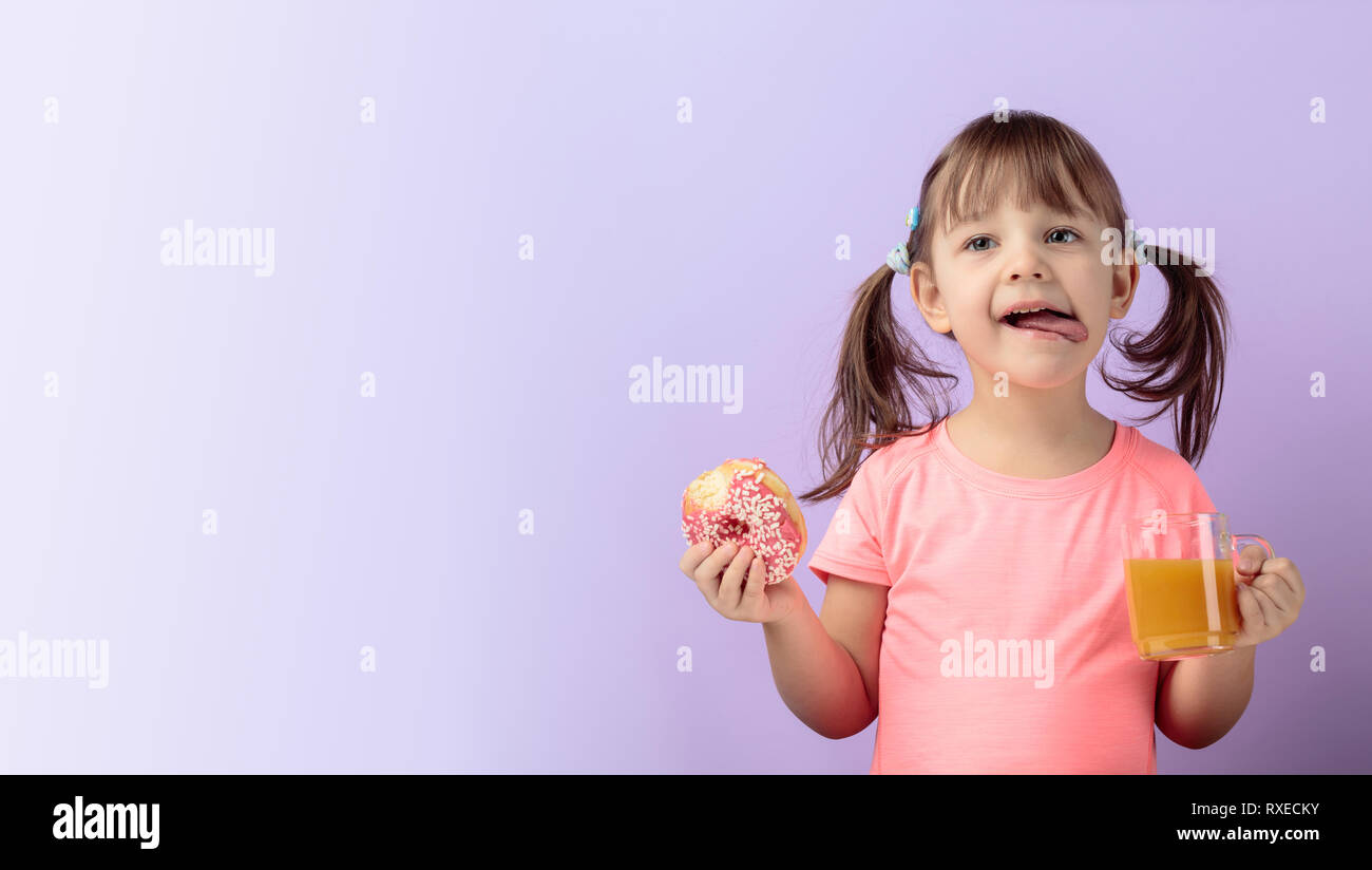 Four-year-old girl in a pink t-shirt eat donut and drink juice. The girl's hair is tied in tails. Purple background. Copy space. Stock Photo