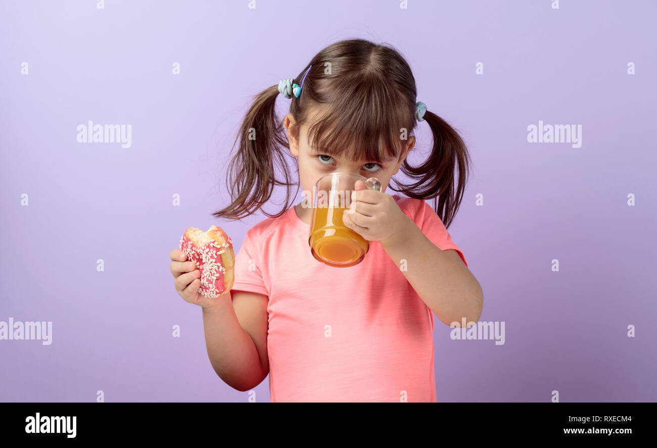 Four-year-old girl in a pink t-shirt eat donut and drink juice. The girl's hair is tied in tails. Purple background. Stock Photo