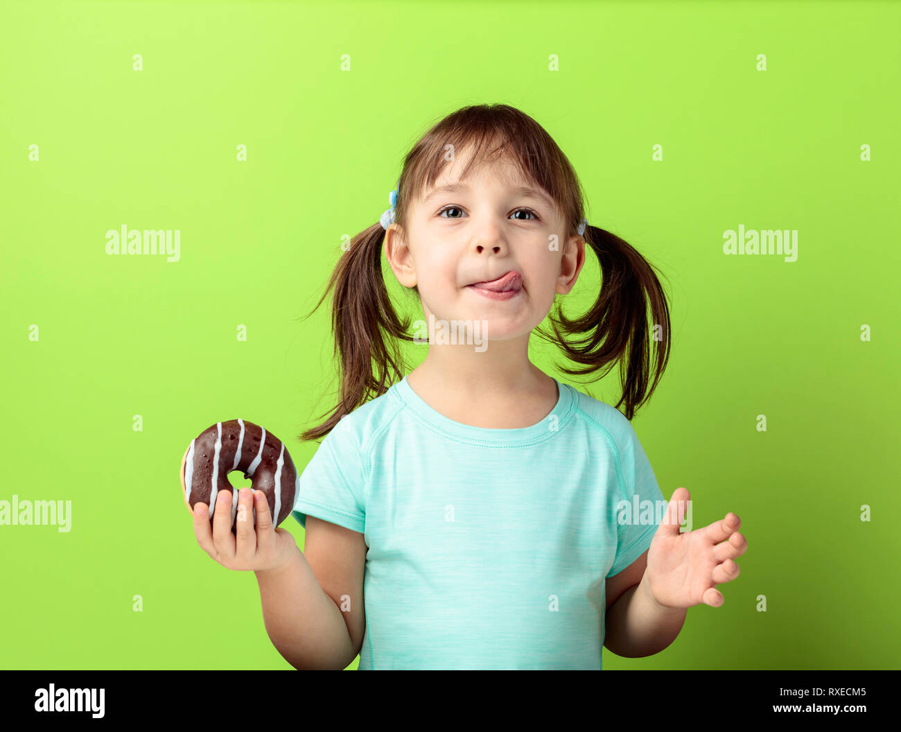 Four-year-old girl in a turquoise t-shirt eat donut. The girl's hair is tied in tails. green background. Stock Photo