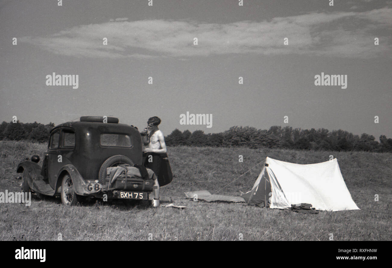 1930s, historical, a young man has a shave standing next to a car of the era (with a GB sticker) parked in a open field next to a small tent, in the Sudentenland, Czechoslovakia. Stock Photo