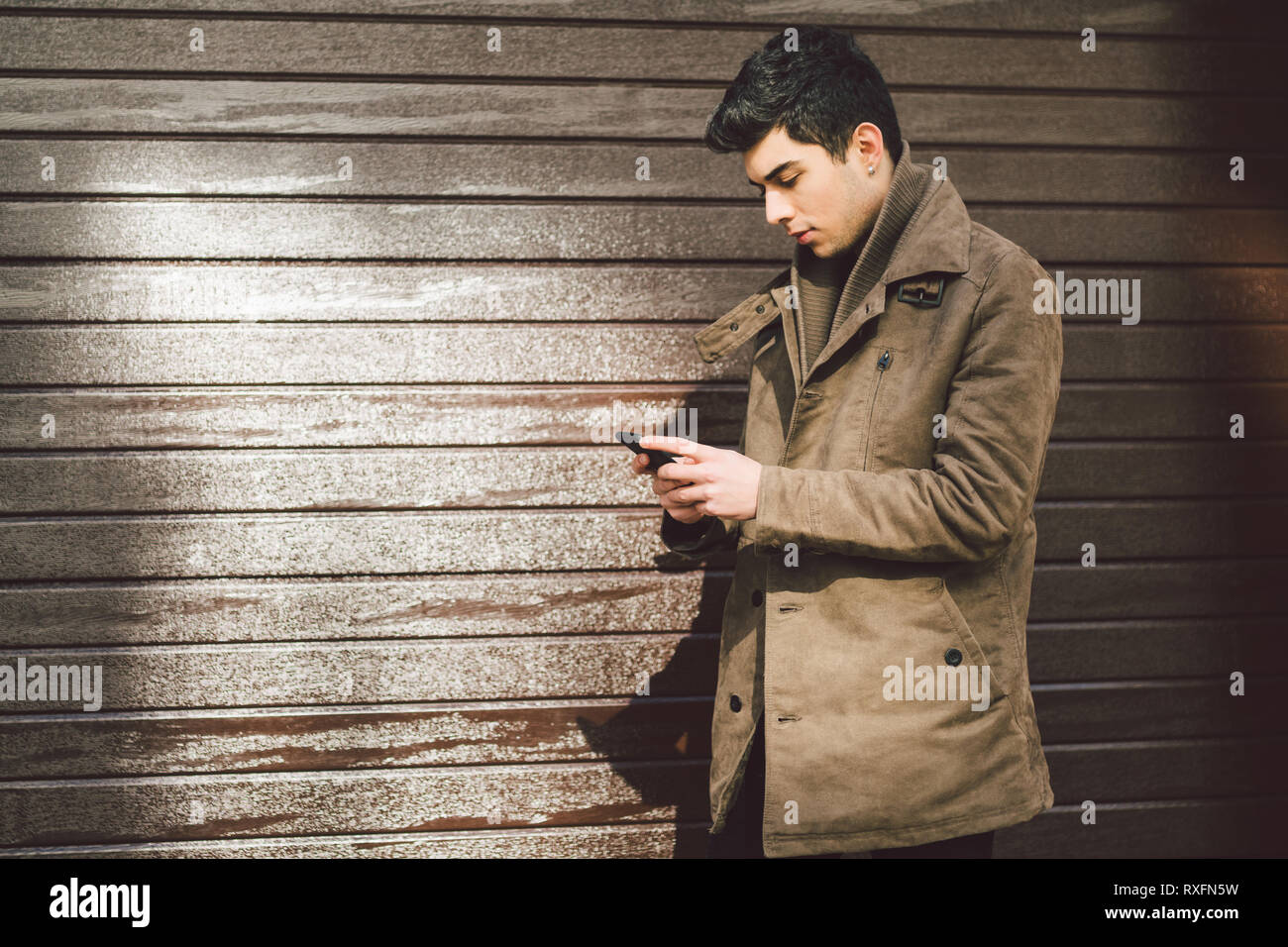 Portrait of young handsome Turkish male model of Mediterranean race brunet in leather coat using hand phone of technology businessman on street in sun Stock Photo