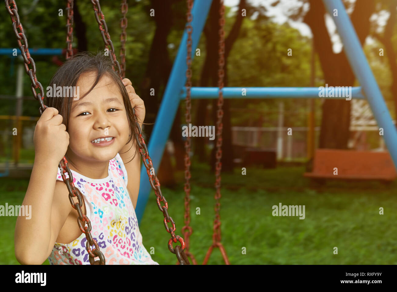Happy smiling asian small girl hang on swings in park background Stock Photo