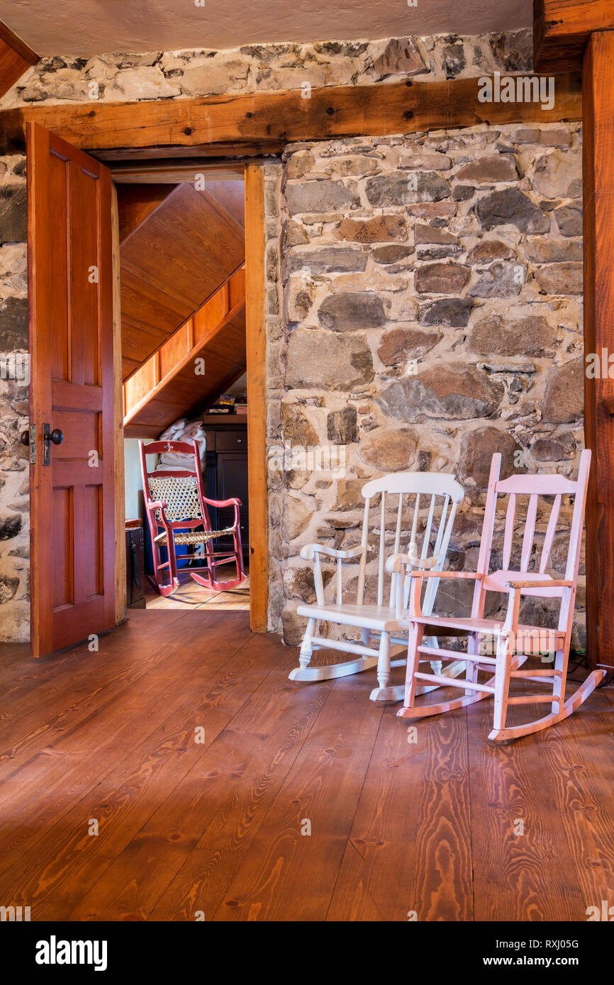 White and pink painted wooden rocking chairs next to opened door of master bedroom with red wood woven rawhide seat chair on upstairs hallway with pinewood floorboards inside an old 1826 cottage style fieldstone house, Quebec, Canada. This image is property released. CUPR0298 Stock Photo