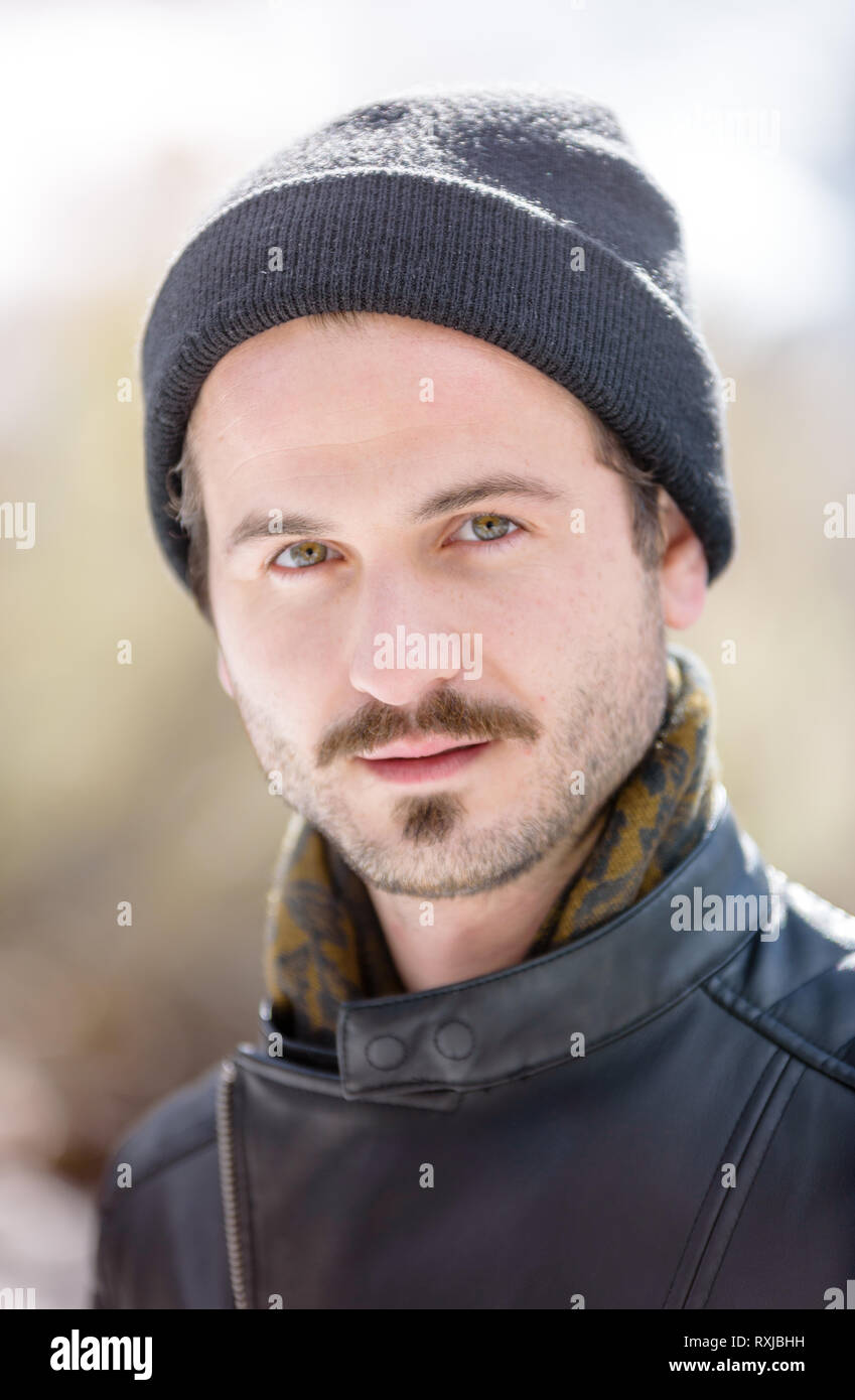 a handsome young man in a leather jacket and beanie posing in the snow covered mountains. Fashion/editorial style image Stock Photo