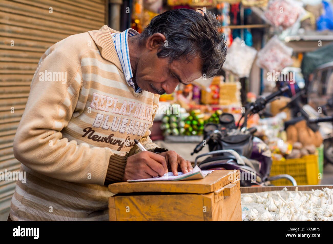 a middle aged Indian street hawker wearing sweater standing, looking down and writing something on a notebook in the afternoon in winter. He is 50-55  Stock Photo