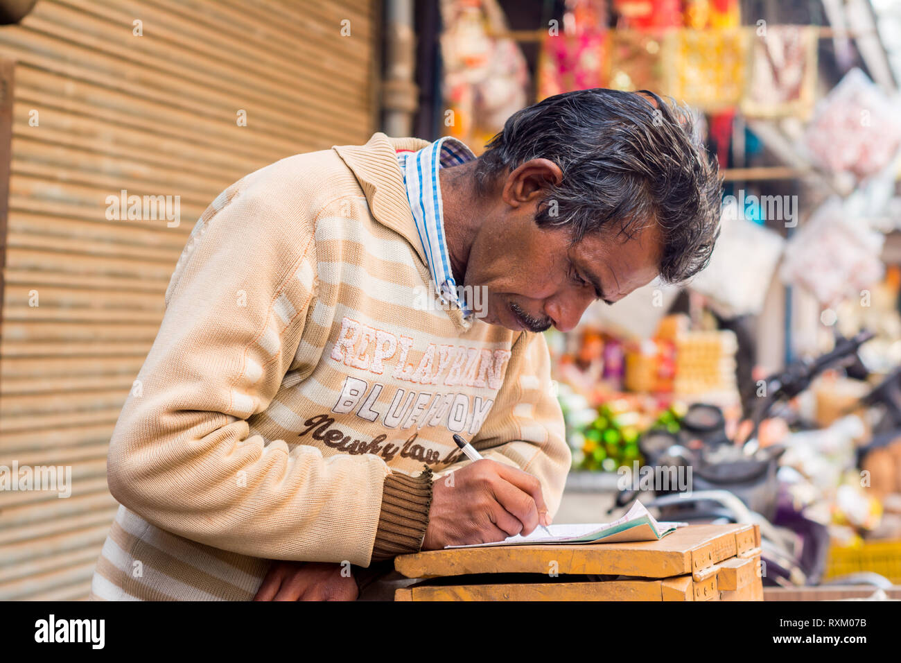 a middle aged Indian man wearing sweater standing, looking down and writing something on a notebook in the afternoon in winter. He is 50-55 years old. Stock Photo