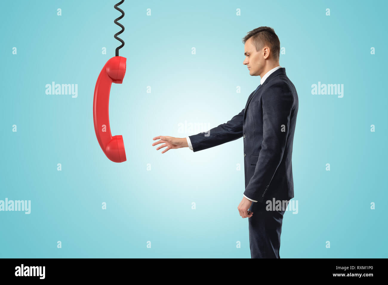 Side view of businessman standing and reaching out for big red landline phone receiver dangling down on wire from above. Stock Photo
