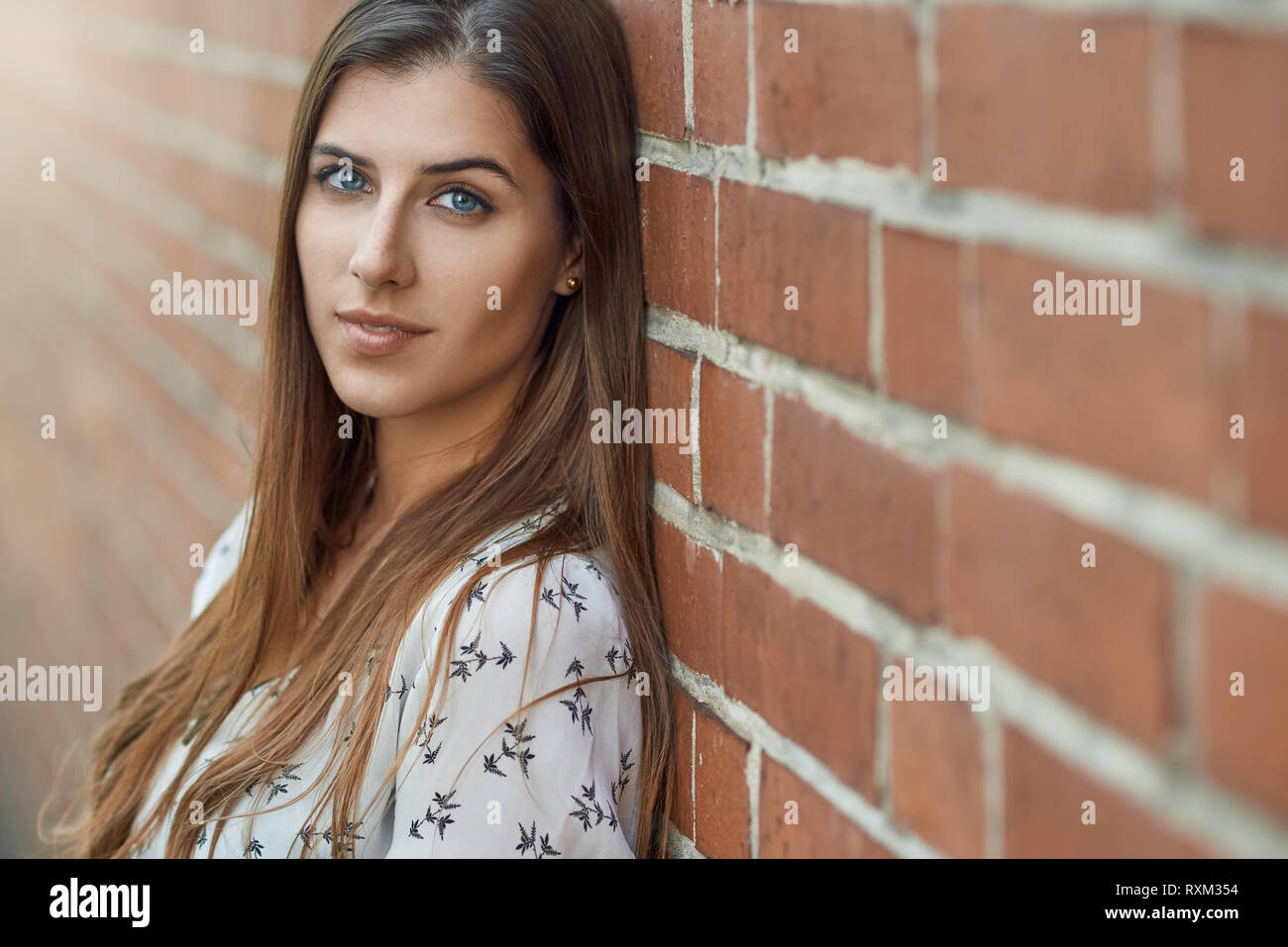 Portrait of young beautiful brunette woman with friendly pretty face, in white blouse, looking at camera while standing near red brick wall with copy  Stock Photo