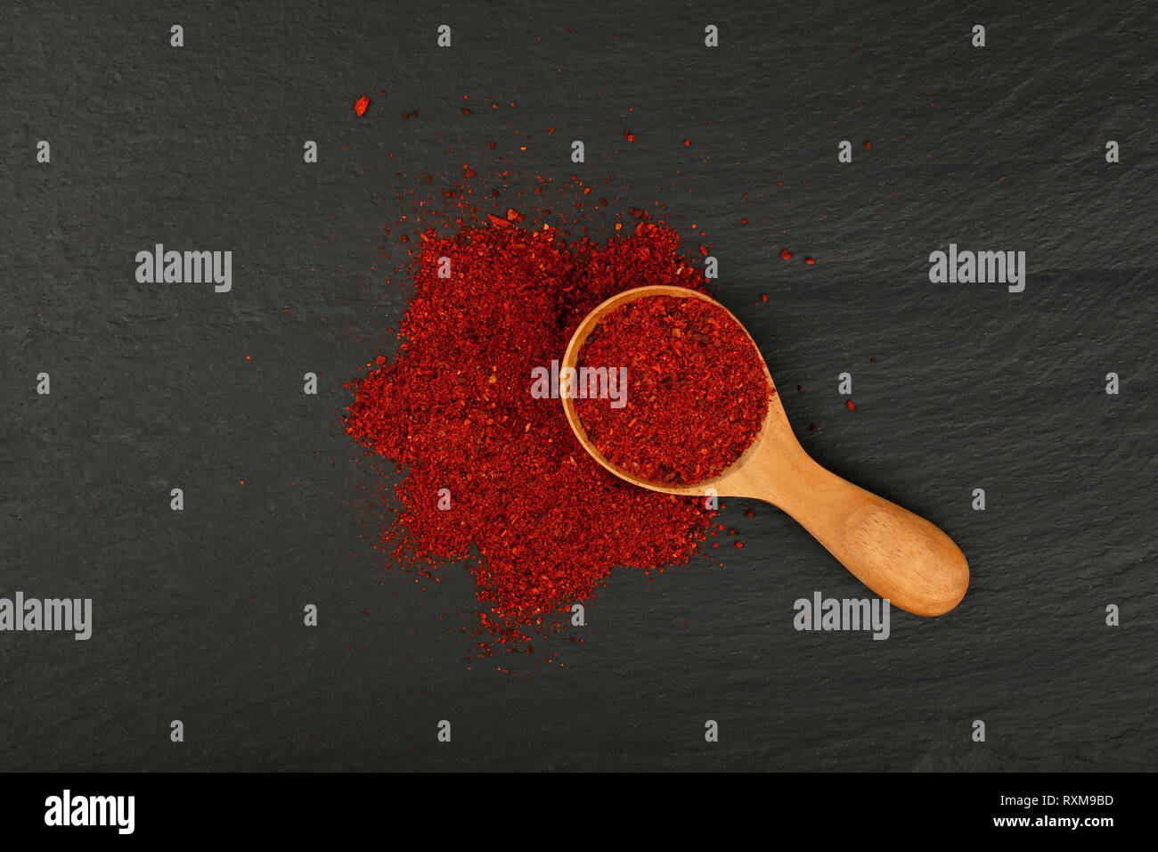 Close up one wooden scoop spoon full of ground sun dried tomato or red chili pepper on background of black slate cooking board, elevated top view, dir Stock Photo