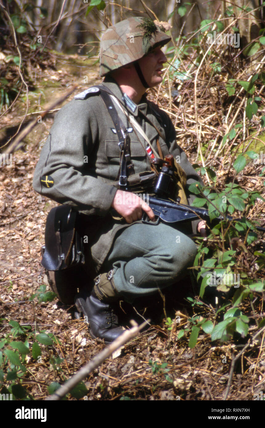 A WW2 German soldier wears period WW2 uniform he kneels in woodland holding a Sten Machine Gun. Stock Photo