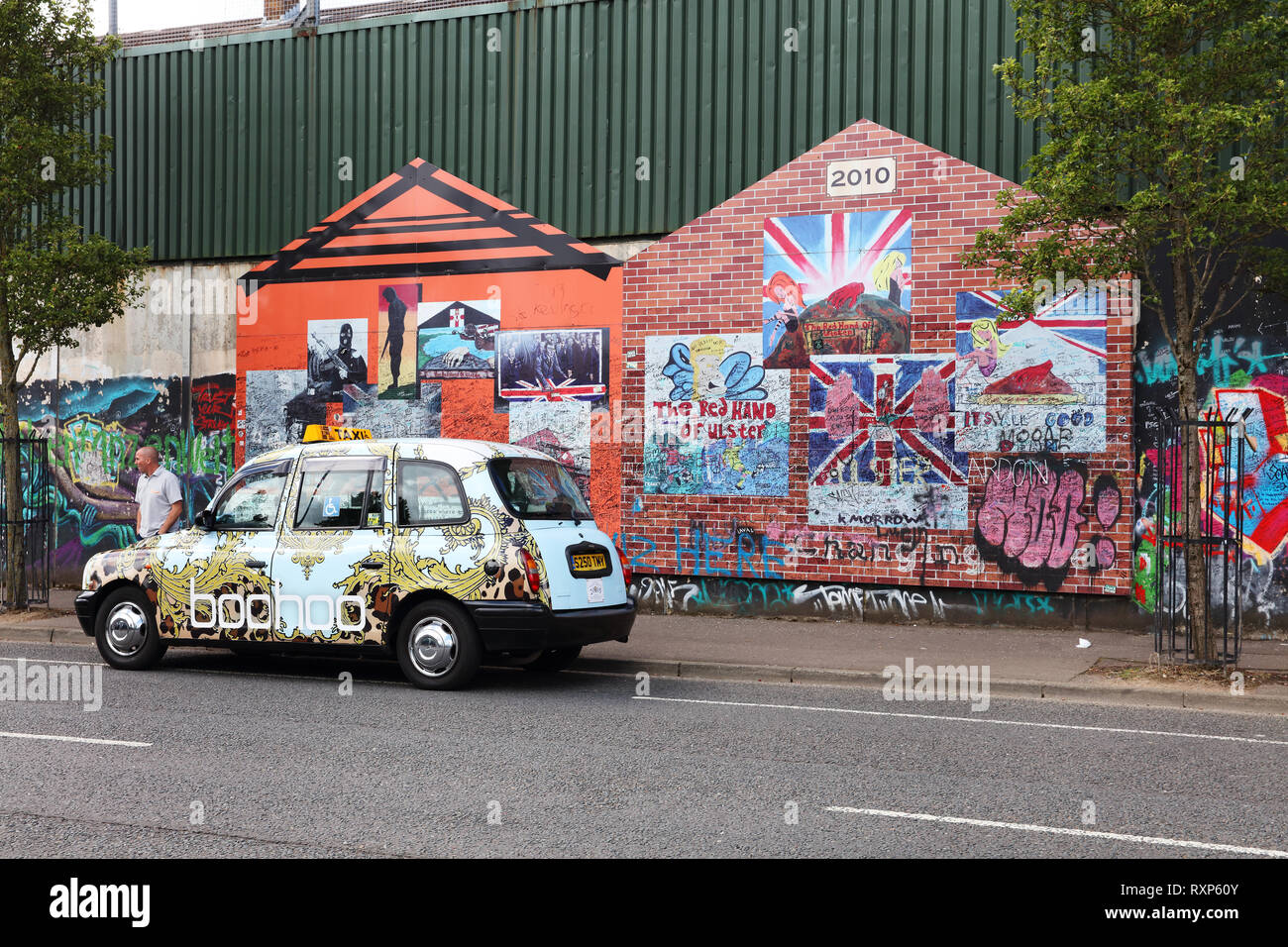 Tour taxi parked in front of the Protestant side of one of several peace walls in Belfast. Tourists are invited to write a thought or sign their names directly over the murals on the wall. Cupar Way, Belfast, Northern Ireland. Stock Photo