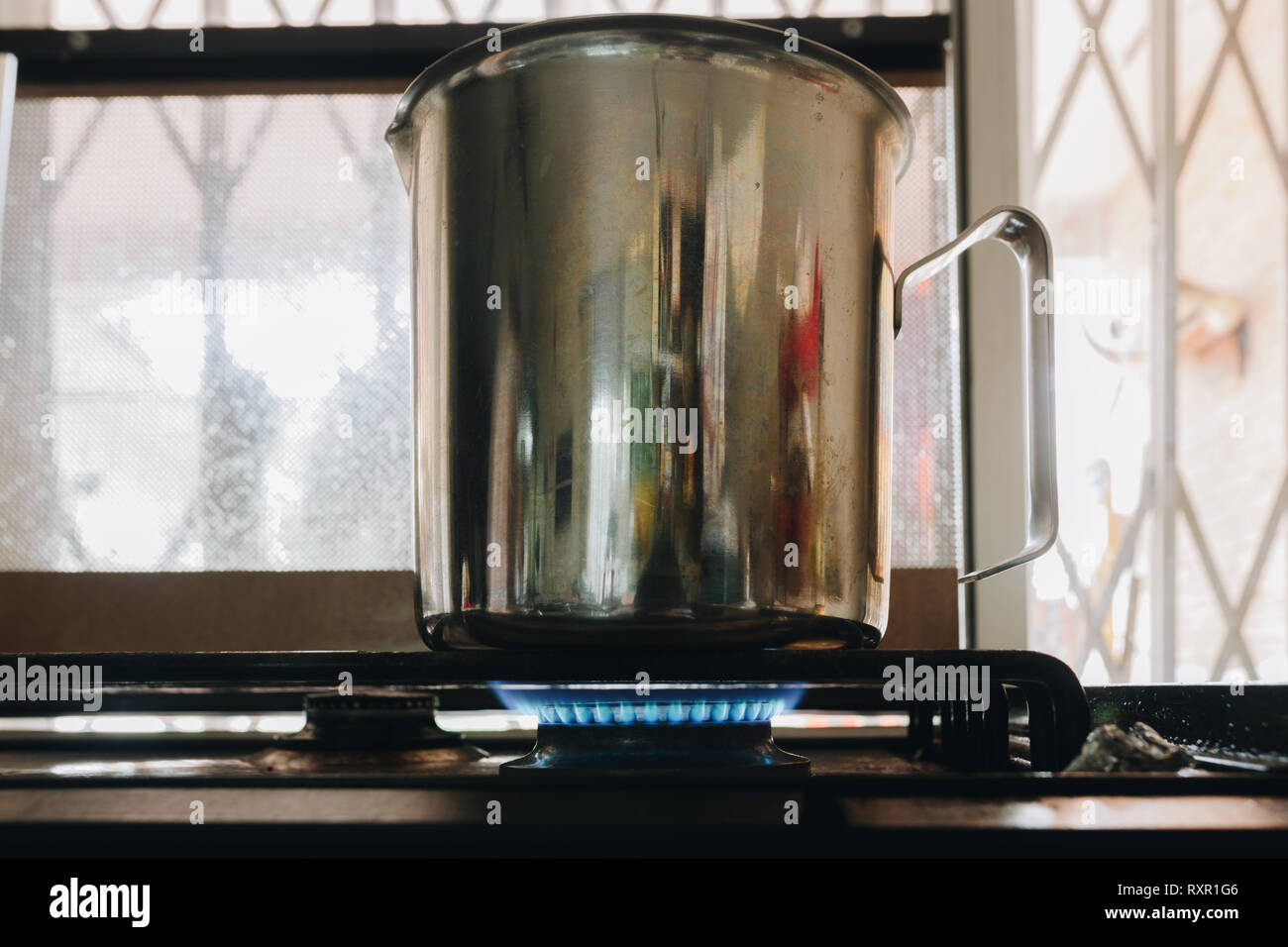 Stainless steel milk kettle, cup, jug on stove. Gas hob cooker flame alight, closeup Stock