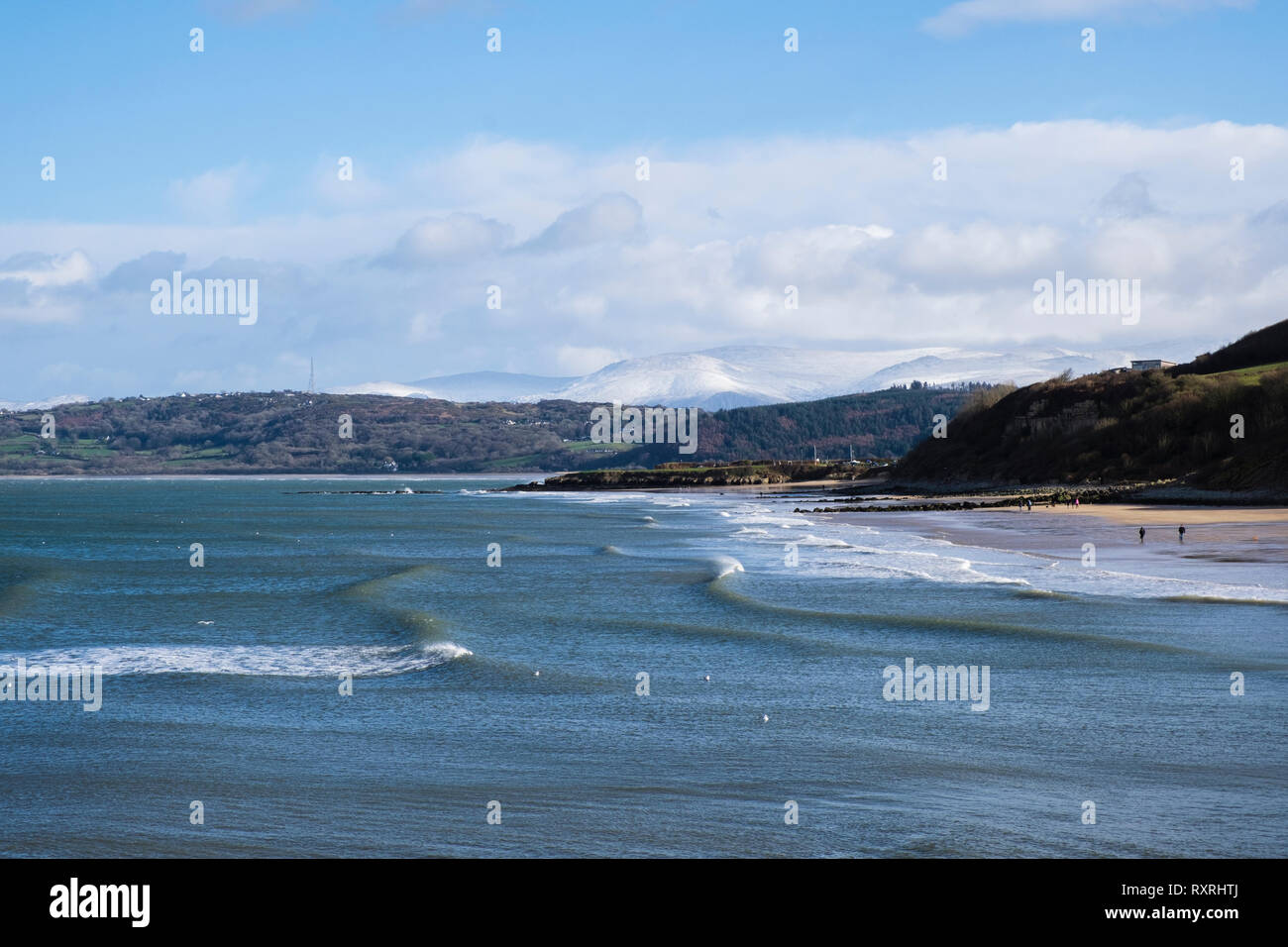 Benllech, Isle of Anglesey, North Wales, UK. 10th Mar 2019. Waves roll on to Benllech beach in sunshine on a cold windy day with snow on distant mountains of Snowdonia. The weekend has seen changeable cold windy weather across the island. A snowstorm followed shortly after. Credit: Realimage/Alamy Live News Stock Photo