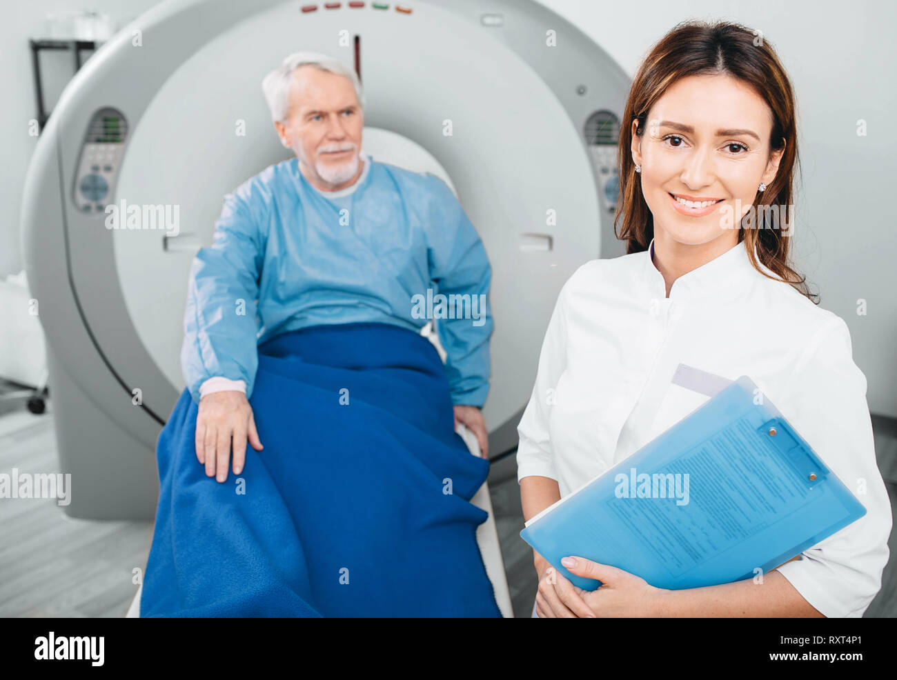Doctor radiologist, smiling while looking at camera. Behind her sitting senior patient. CT scanner in hospital Stock Photo