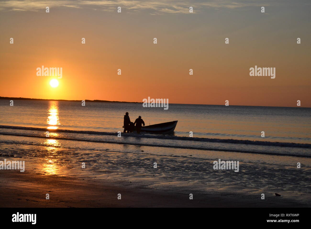 Fishing boat and 2 fishermen setting off, as the sun rises over Filey Brigg Yorkshire UK Stock Photo