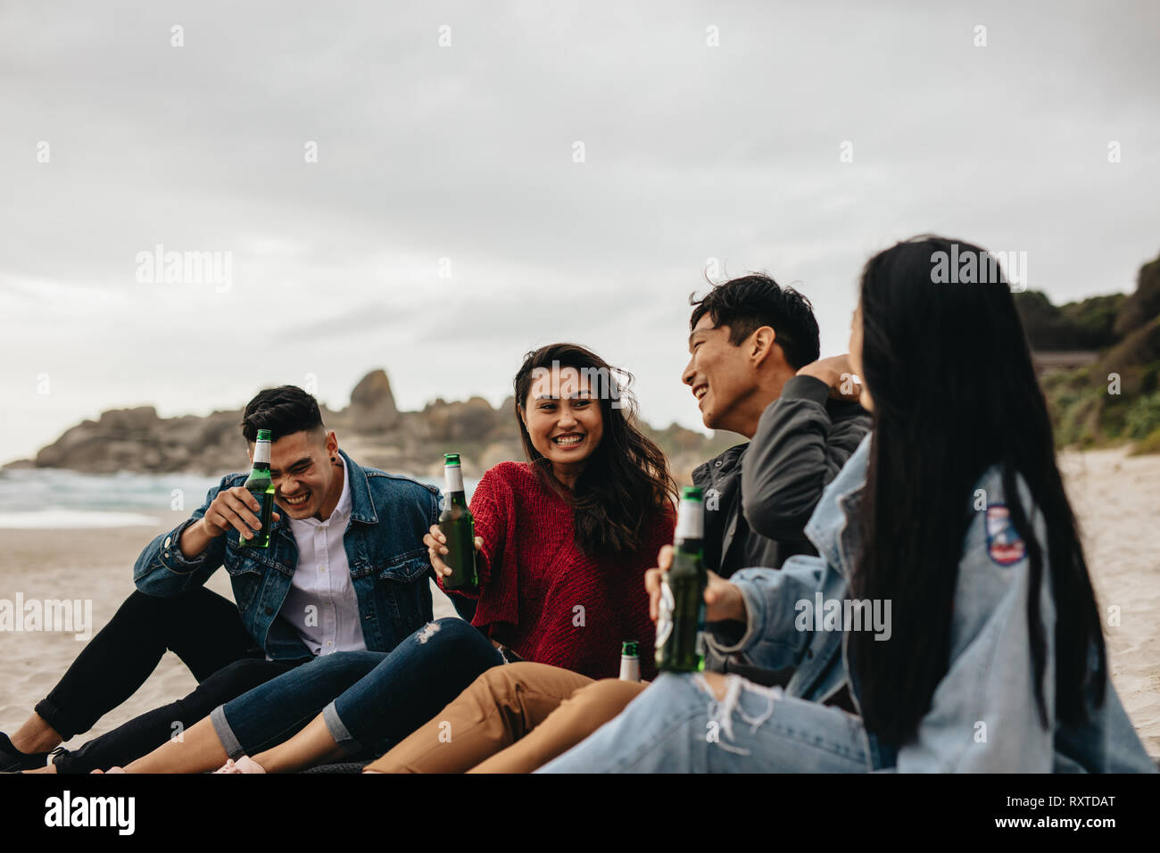 Cheerful young asian men and women sitting at the beach having beers. Two couple partying on the beach. Stock Photo