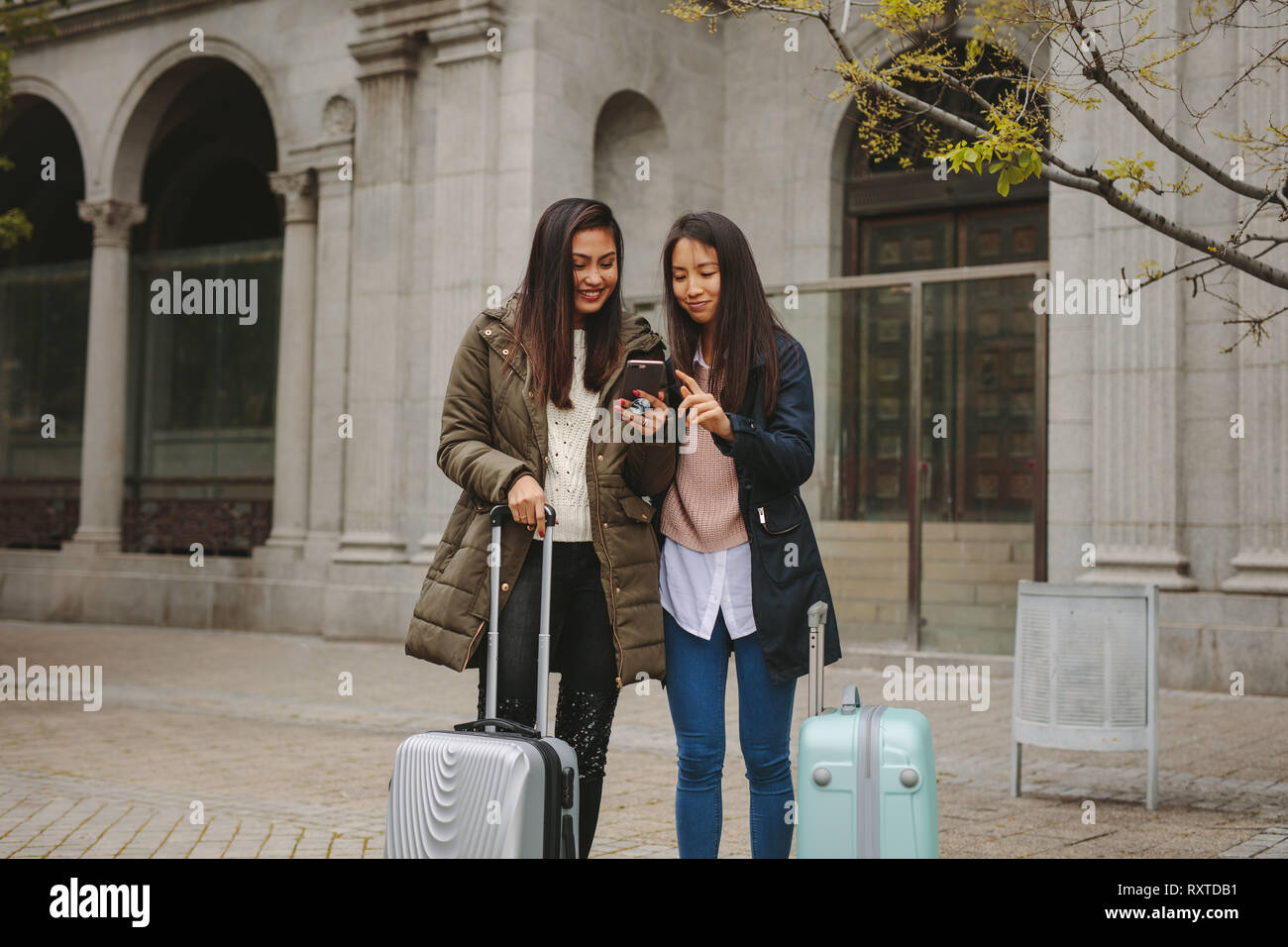 Tourist women standing in street with luggage bags looking for directions in cell phone. Two smiling asian tourists looking at mobile phone. Stock Photo