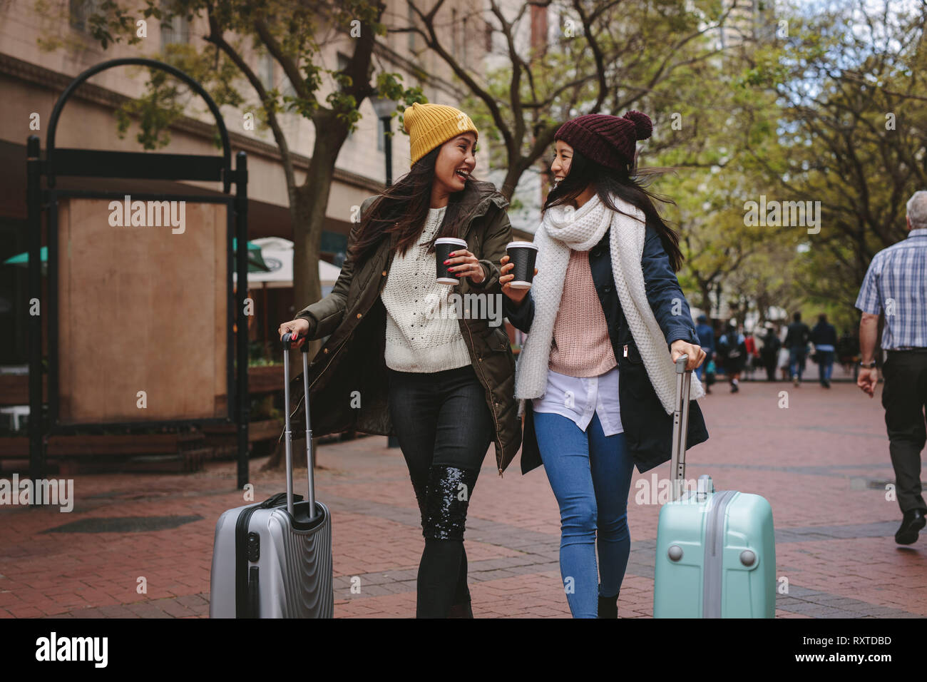 Two smiling woman tourists walking around the city drinking coffee. Two asian women walking on street with luggage bags enjoying coffee. Stock Photo