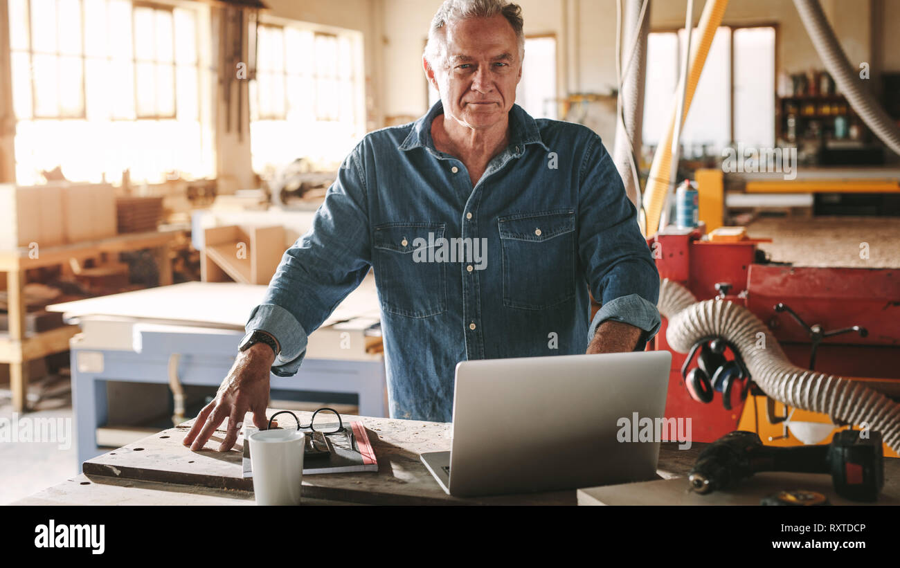 Portrait of confident senior male carpenter standing by his workbench looking at camera. Mature male carpenter in his workshop with laptop on table. Stock Photo