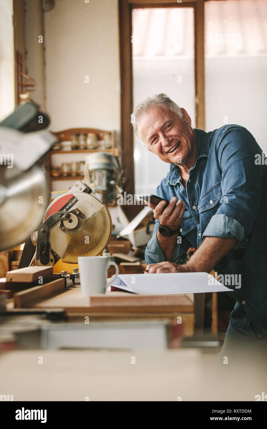 Happy senior man standing by a table in his carpentry workshop with a phone in hand. Mature male carpenter looking at camera and smiling. Stock Photo