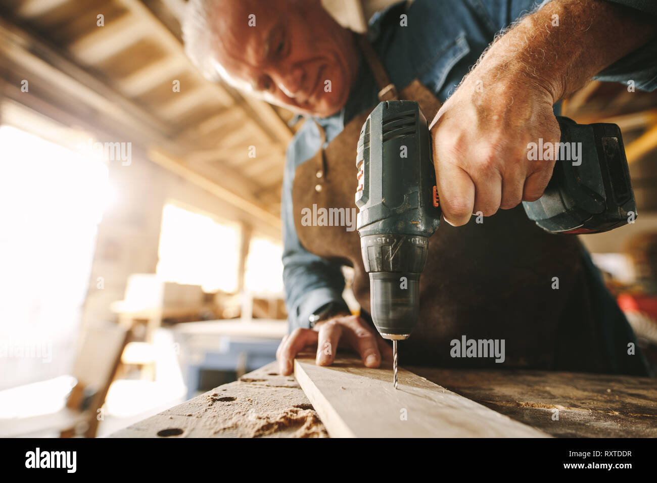 Senior male carpenter drilling a hole in wooden plank on workbench. Focus on carpenter hand preparing furniture. Stock Photo