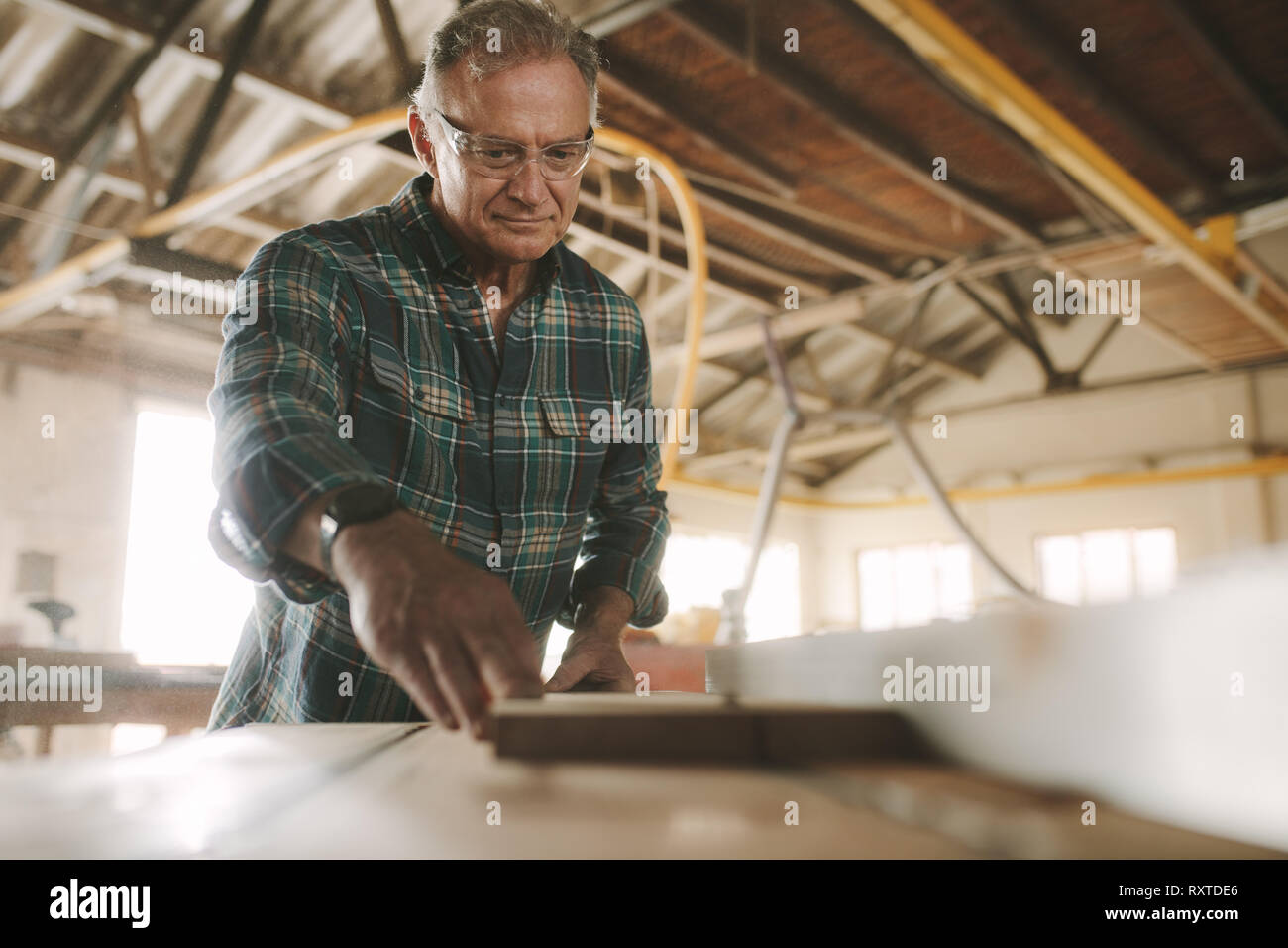 Senior male carpenter working on electric table saw machine cutting wood planks. Skilled mature carpenter working in carpentry workshop. Stock Photo