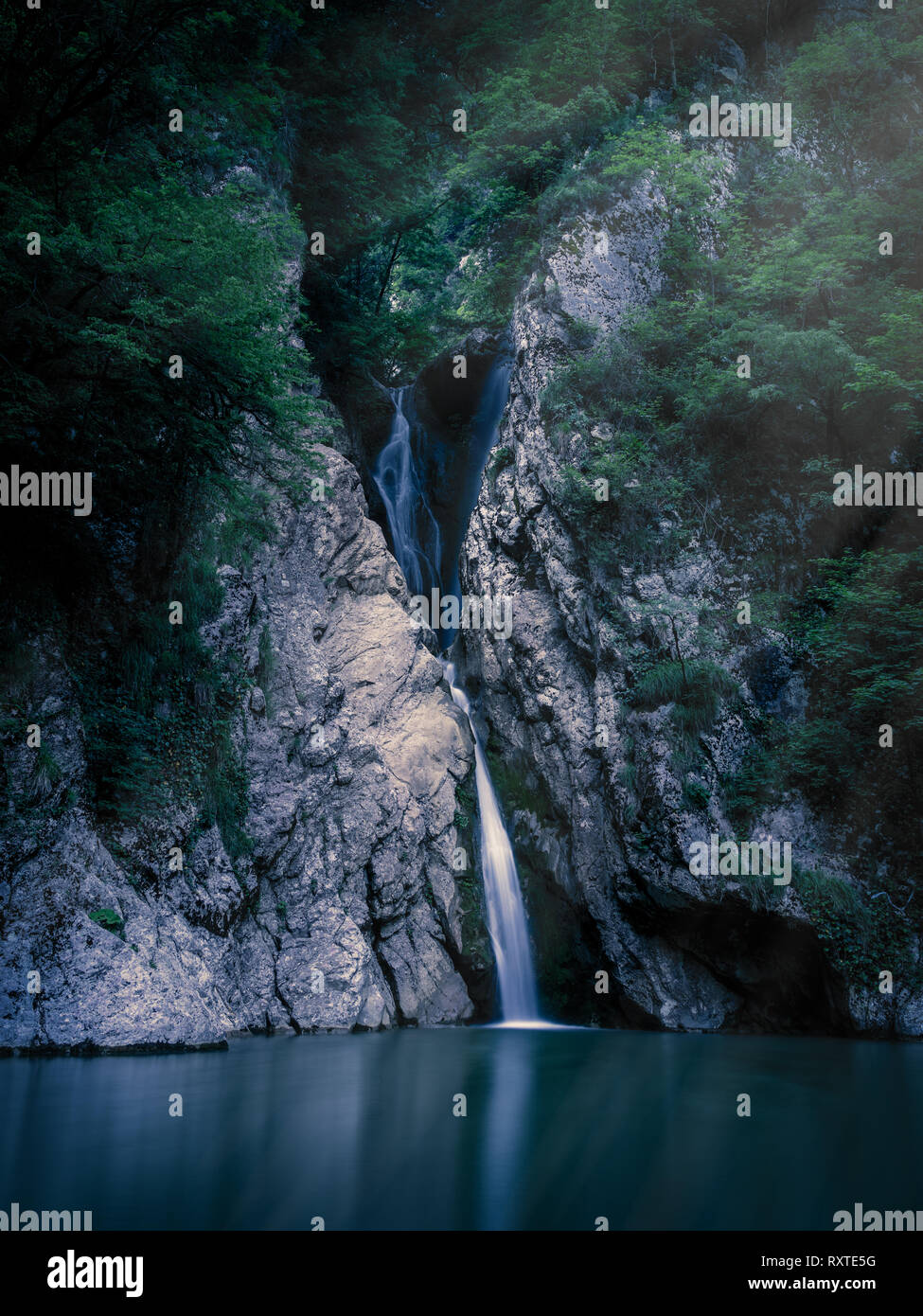 A tall waterfall falls from a cliff into a clear lake at night, lit by the  light of the moon. The waterfall is surrounded by green forest. River Agura  Stock Photo -