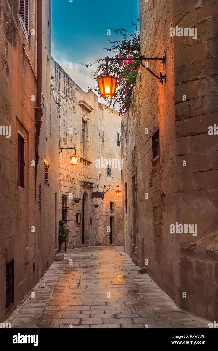 Mdina, Malta: narrow street paved with setts in medieval town with limestone walls and lantern lights in the early evening Stock Photo