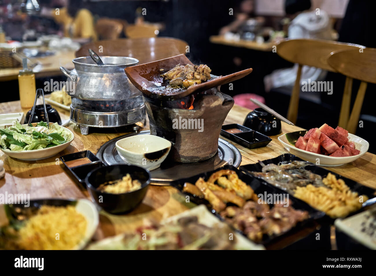Tasty chicken meat is cooking on the hot pot with burning coal on the wooden table in the vietnamese cafe. Varied food on the plates are around it. Cl Stock Photo