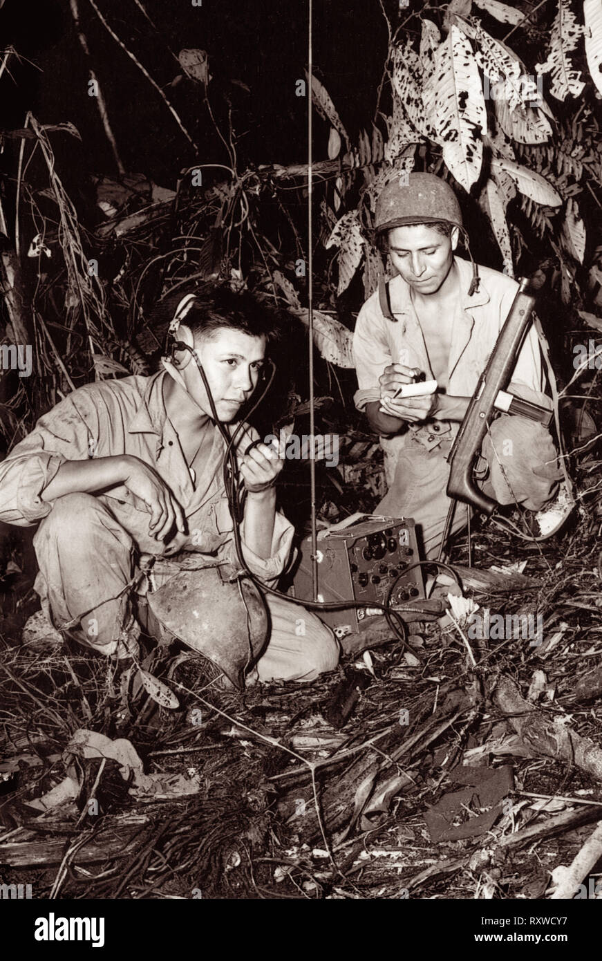 Navajo code-talkers Corporal Henry Bake, Jr., (left) and Private First Class George H. Kirk (right), serving with a Marine Signal Unit, operate a portable radio set in a clearing they've just hacked in the dense jungle close behind the front lines on Bougainville Island in Papua New Guinea during World War II in December 1943. Stock Photo