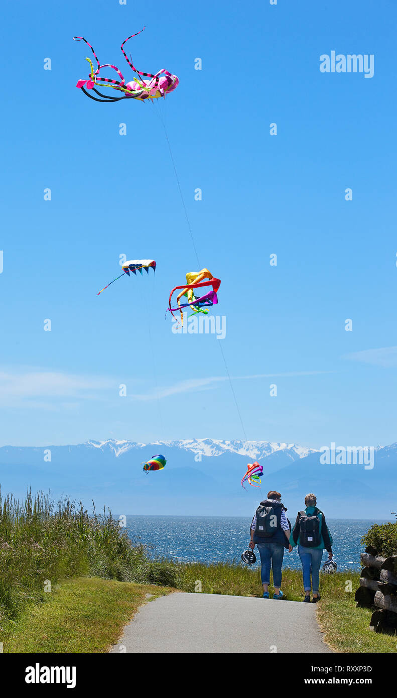 Elaborate kites flying high above hikers on the shoreline trail at Holland Point Park, Victoria, British Columbia, Canada Stock Photo