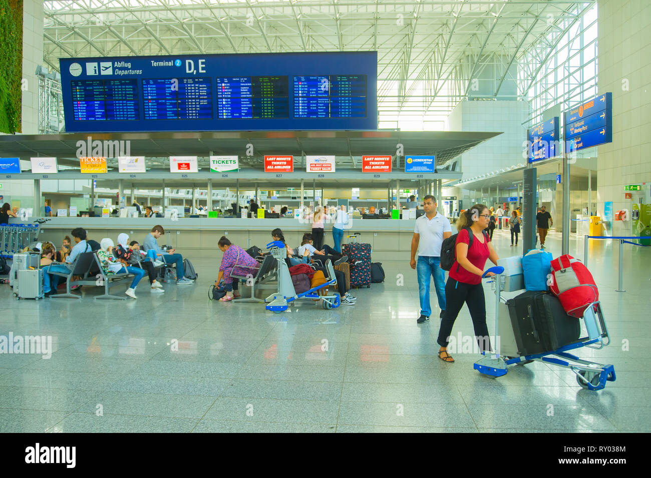 FRANKFURT AM MAIN, GERMANY - AUGUST 29, 2018: Frankfurt international airport terminal, woman with luggage cart, passengers waiting on chairs under ti Stock Photo