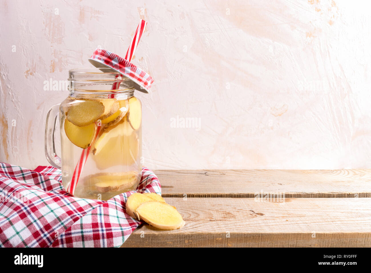 Glass jar with ginger water with drinking straw and tartan cap and cloth on wooden planks on light background. Stock Photo