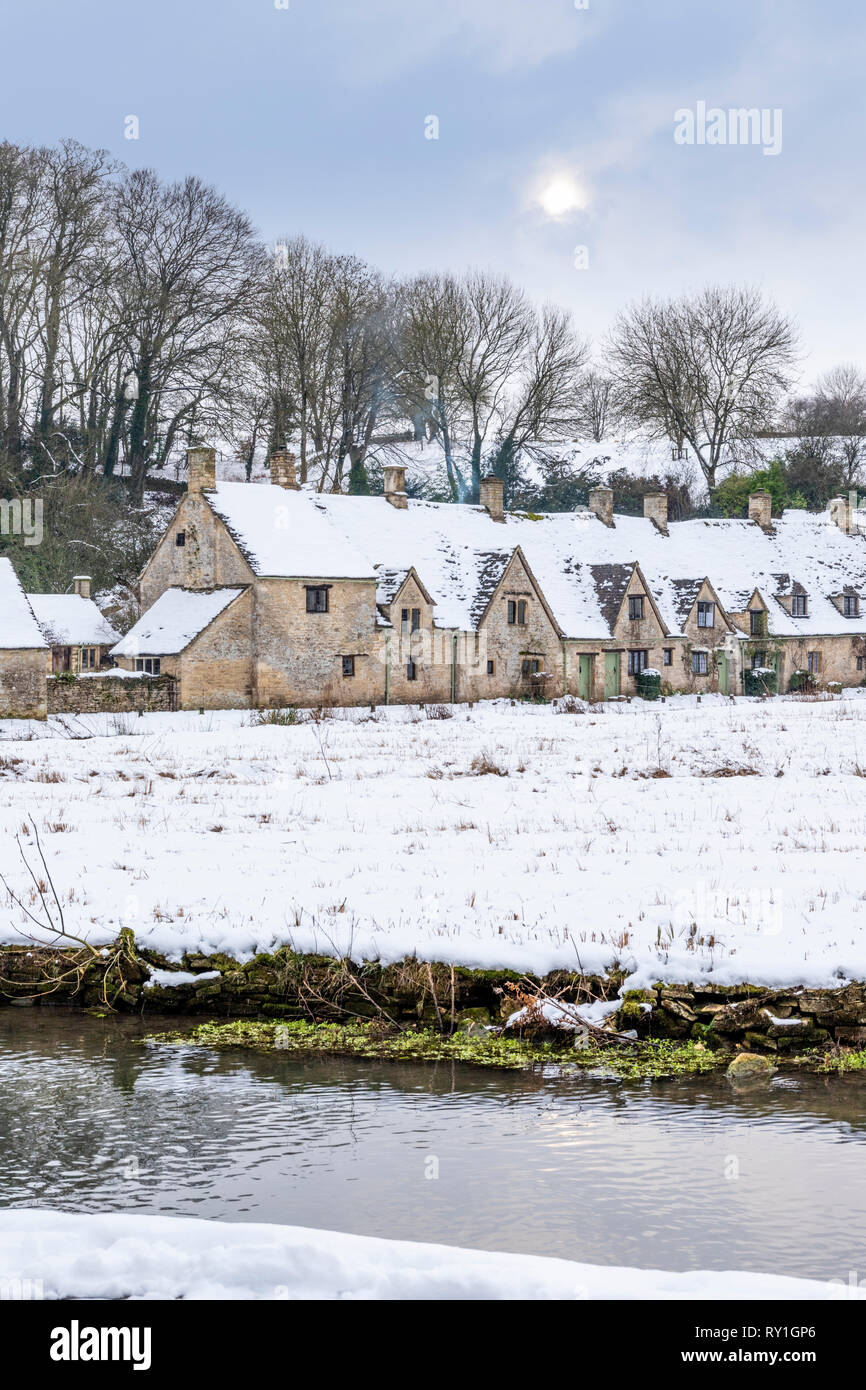 Arlington Row covered with snow beside the River Coln under a pale wintry sky at Bibury, Gloucestershire UK Stock Photo