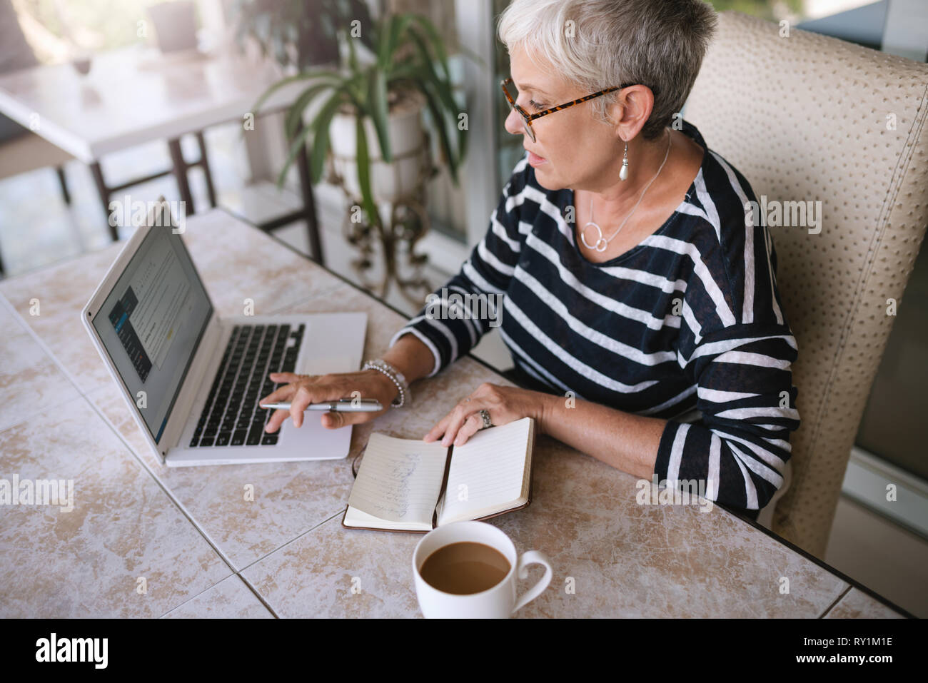 Mature beautiful woman working on her laptop at home. Elderly woman typing on her laptop while reading notes from her notebook outside on the balcony Stock Photo