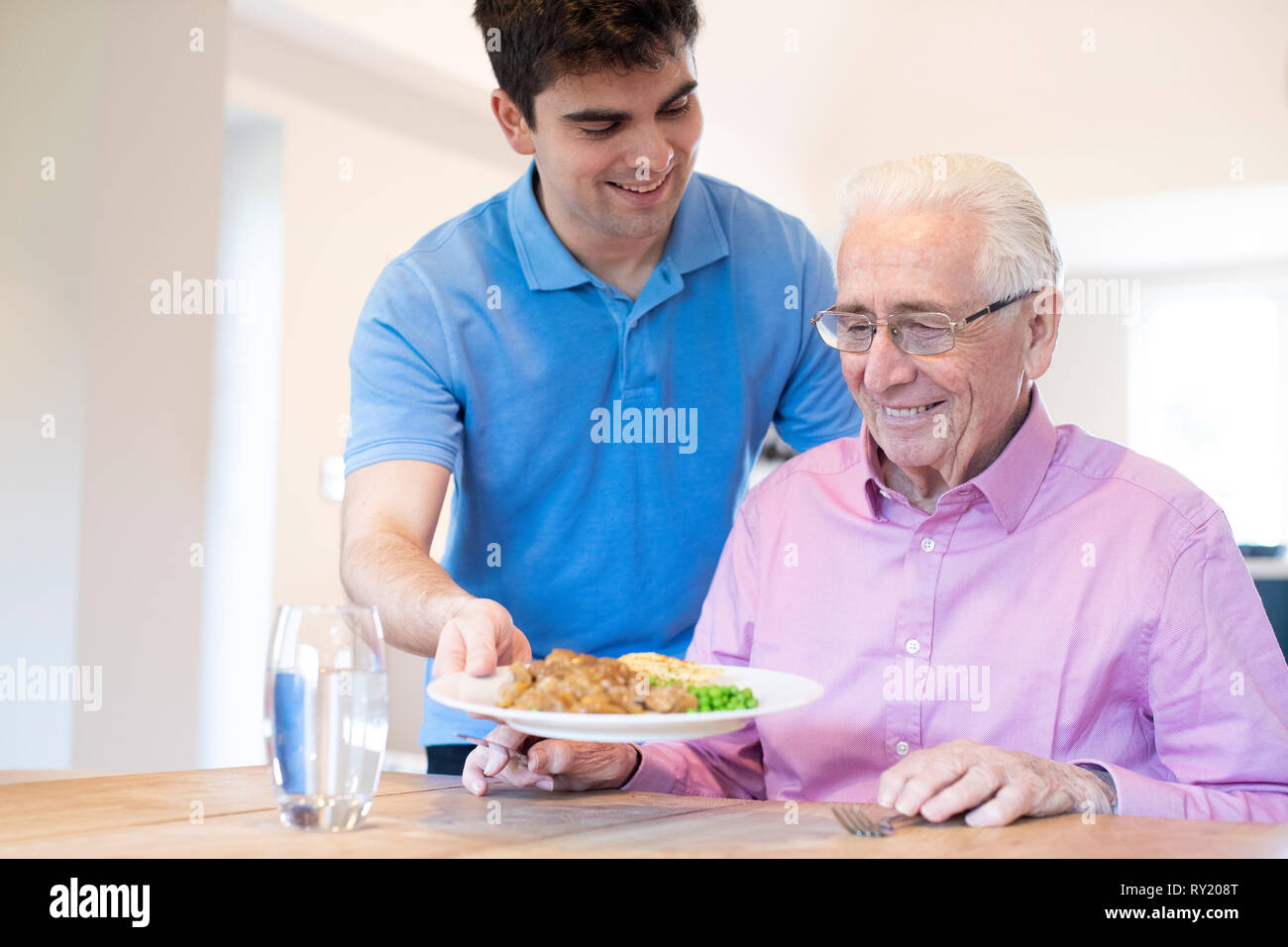 Male Care Assistant Serving Meal To Senior Male Seated At Table Stock Photo
