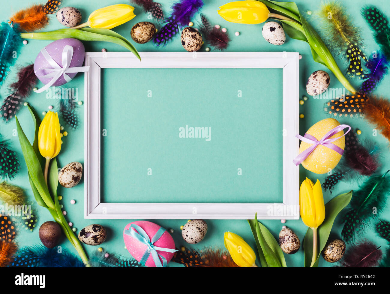 Easter Frame of yellow tulips, decorated eggs and feathers on turquoise ...